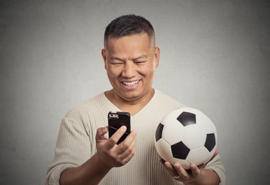 Portrait excited smiling surprised man looking on cell smart phone watching game holding football isolated grey wall background. Positive human emotion, face expression. Data plan online gaming